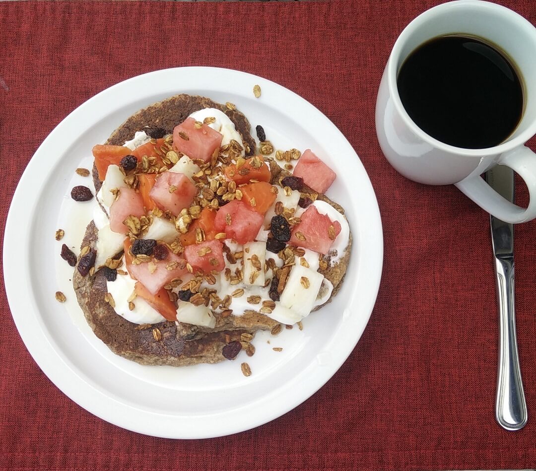 A from-scratch pancake topped with yogurt, watermelon, banana and granola, with a cup of black coffee on a red cloth